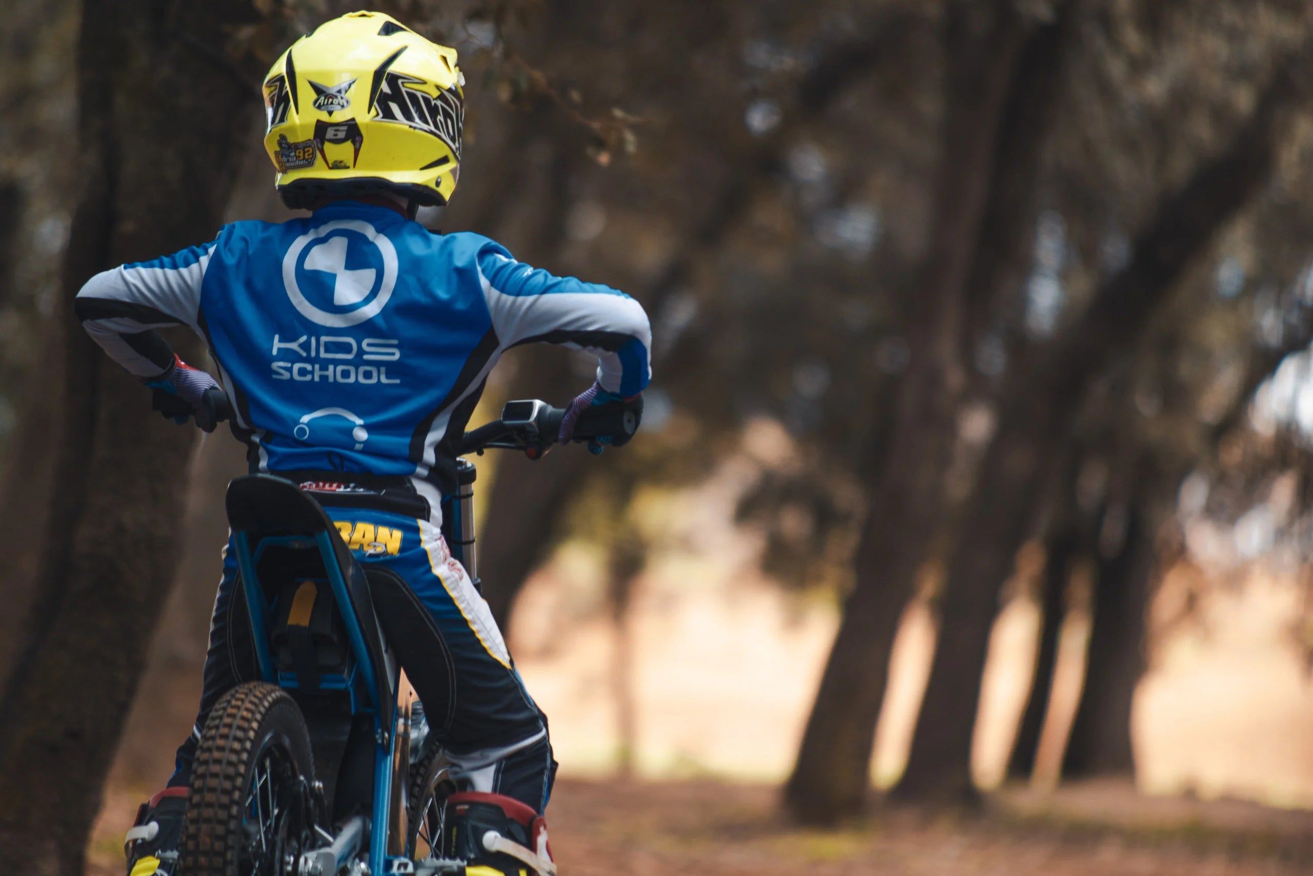 Child on a balance bike with a helmet and 'Kids School' shirt in a forest setting