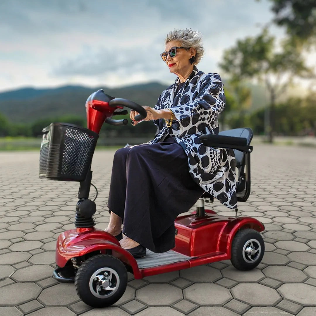 Woman riding a red mobility scooter outdoors on a paved path with mountains in the background.