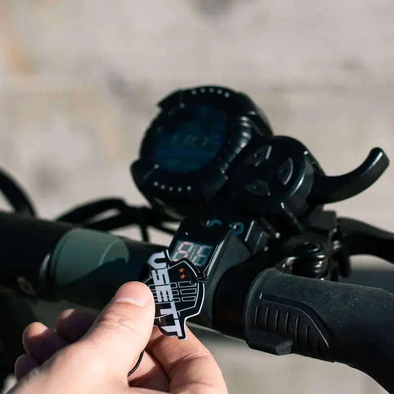Close-up of motorcycle handlebar controls with a hand holding a 'DSEI-TEC' sticker.