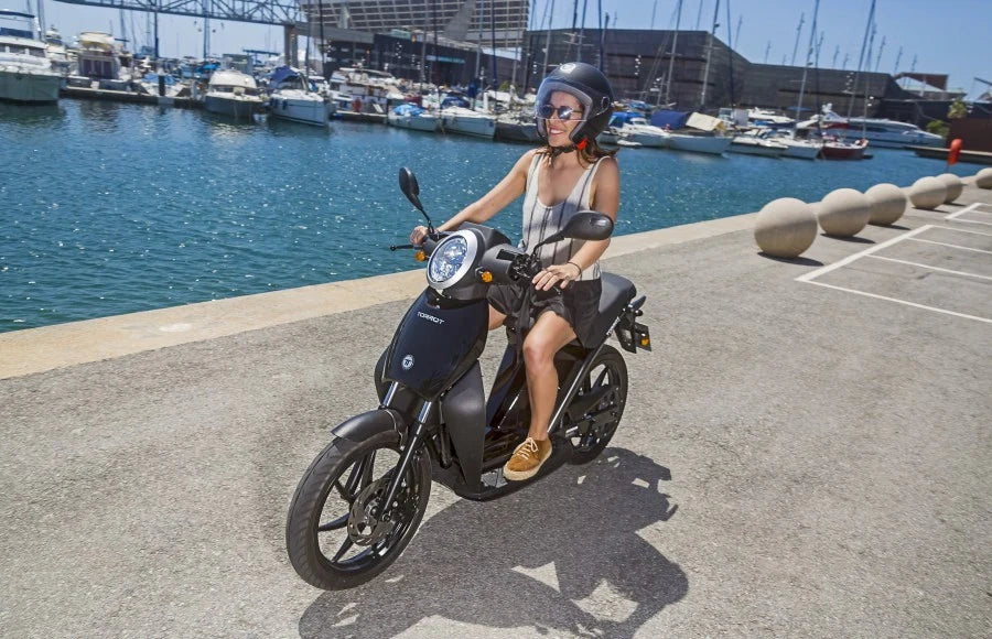 Person riding a scooter by a waterfront with boats in the background