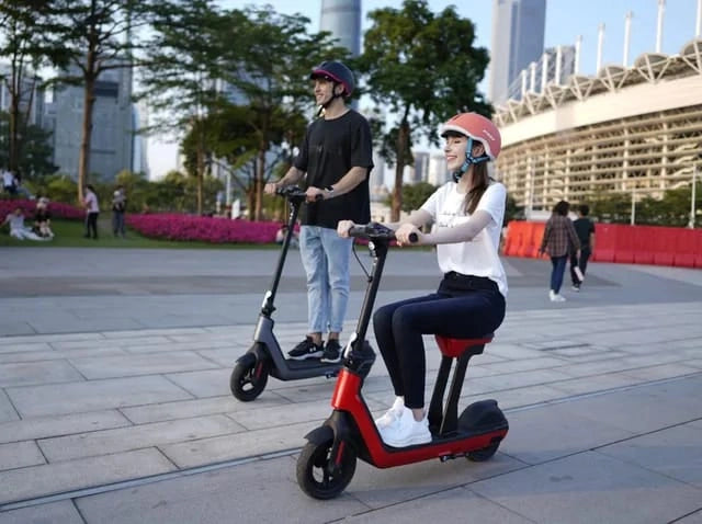 Two people riding electric scooters in an urban setting with buildings and greenery in the background.