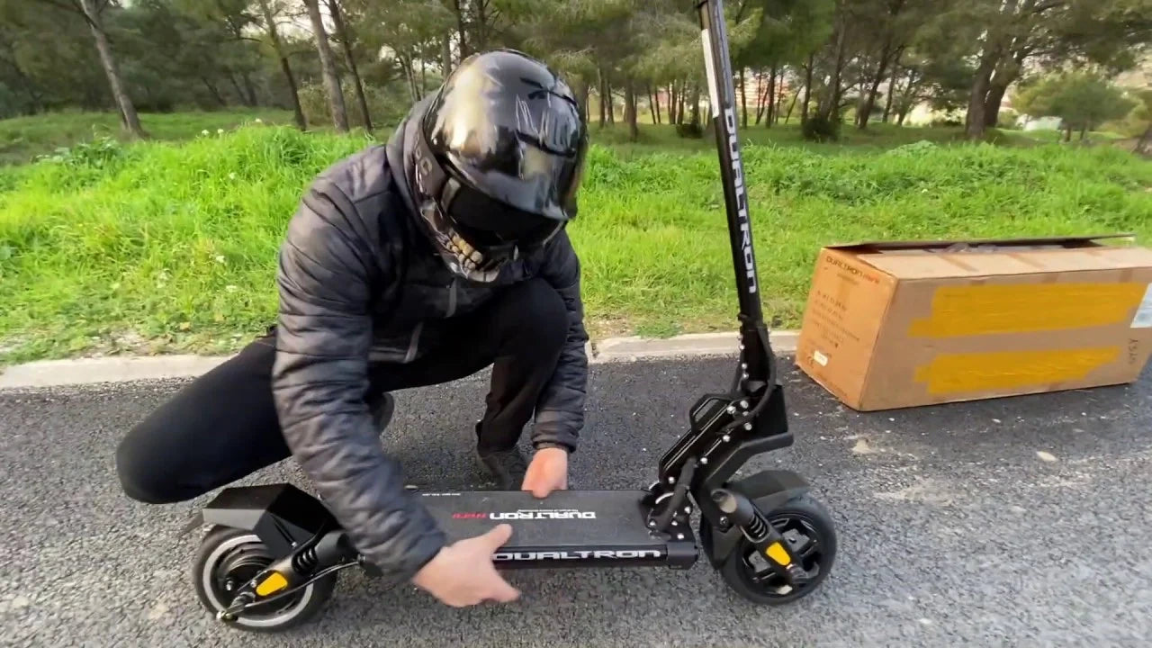 Person wearing a helmet and dark clothing interacting with a black electric scooter on a paved path with grass and trees in the background.