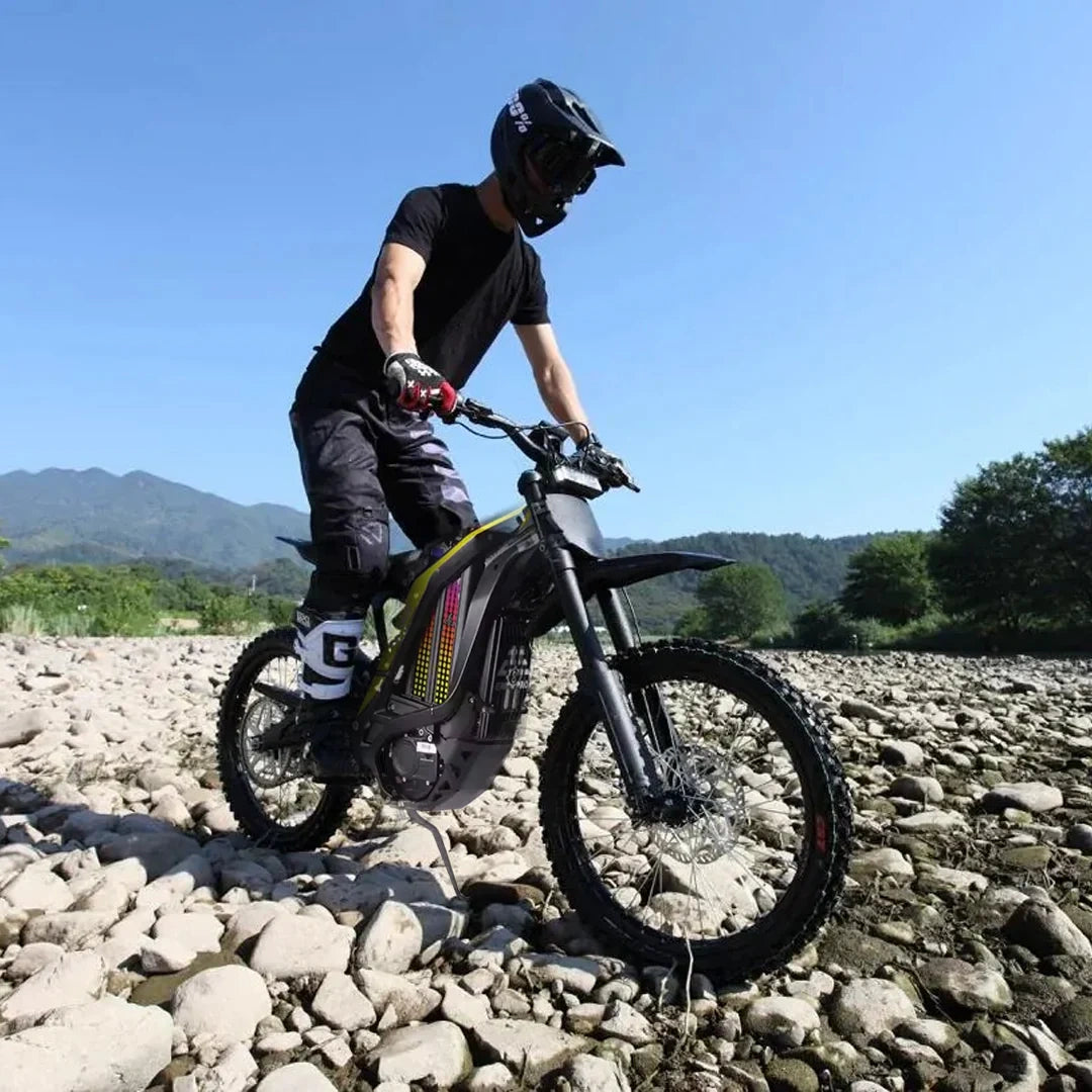 Person riding a dirt bike on rocky terrain with a clear blue sky and greenery in the background