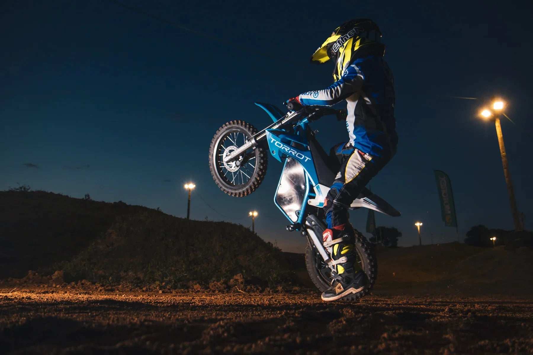 Motorcyclist performing a stunt on a dirt track at night with lights illuminating the scene.
