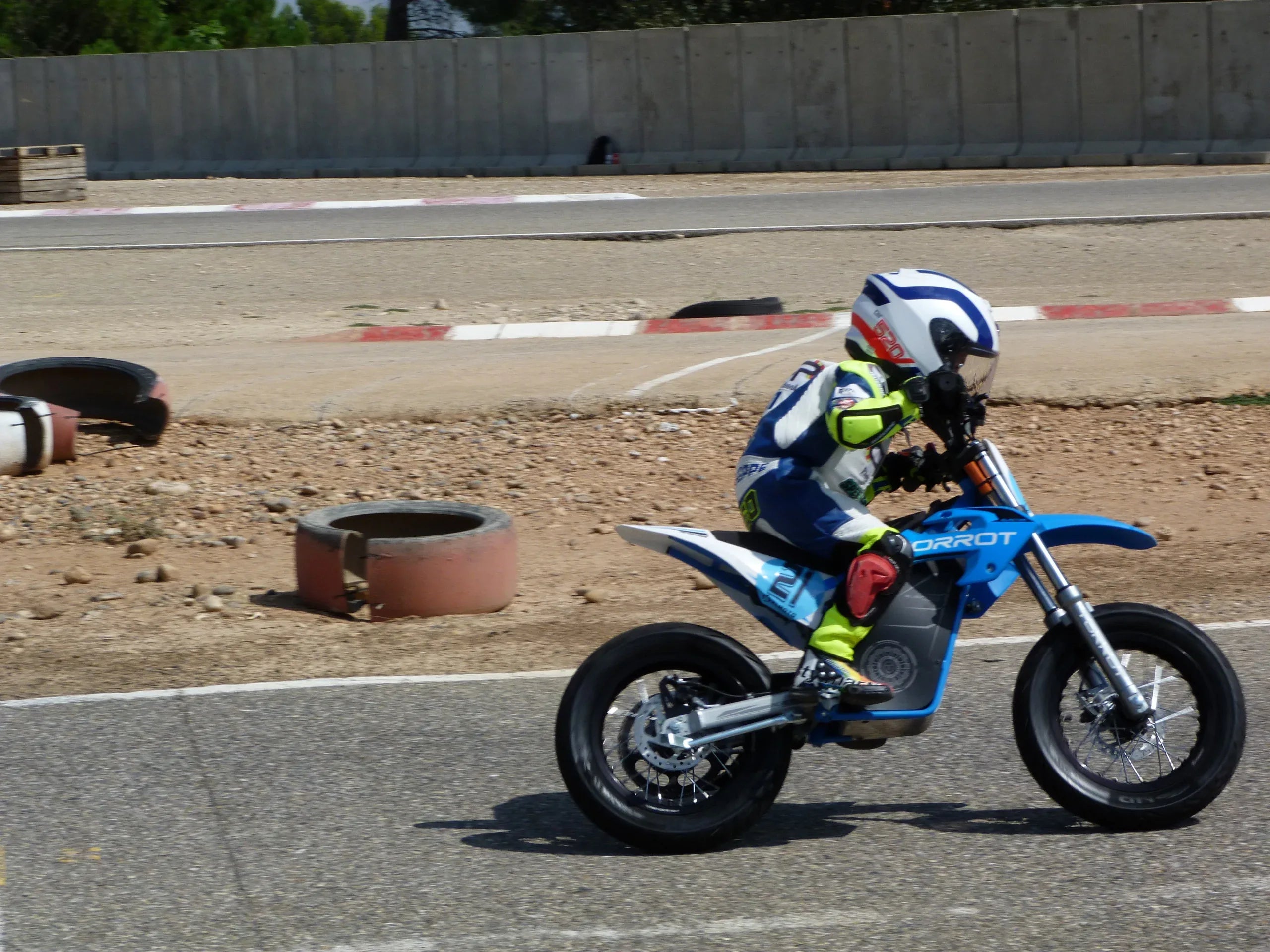 Child on a blue motorcycle at a race track with tires and barriers in the background.