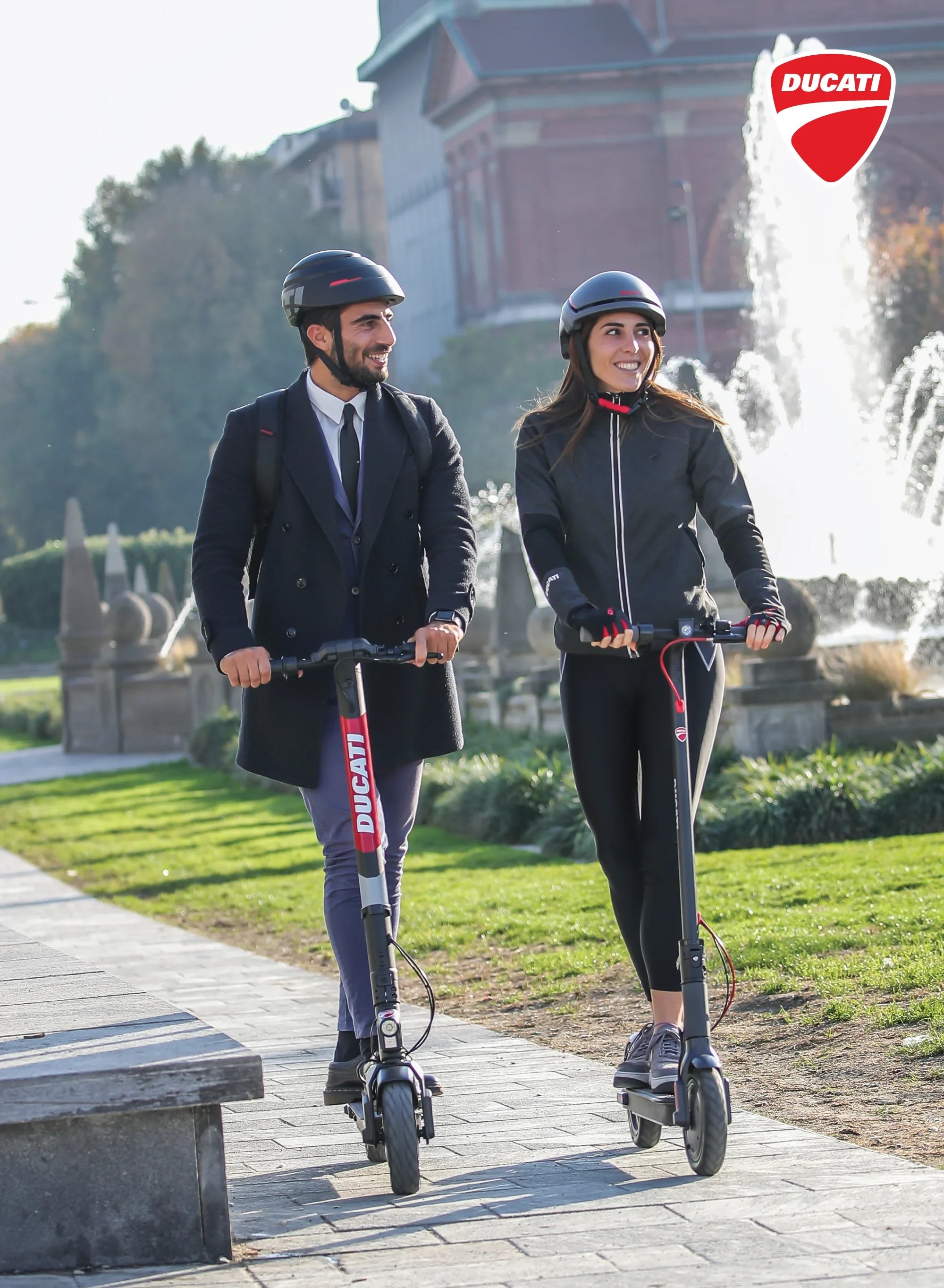Two people riding Ducati electric scooters in a park with a fountain in the background.