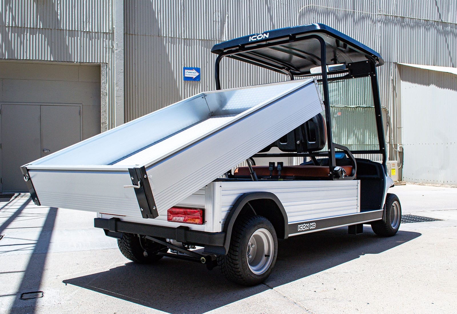ocean cartz rear three-quarter view of a white utility electric vehicle with a fully raised dump bed, demonstrating its unloading capability. This specialized electric work cart features a silver aluminum cargo bed, black roof, and a low profile. The dump cart is parked in an industrial area, highlighting its heavy-duty design for commercial hauling and utility operations.