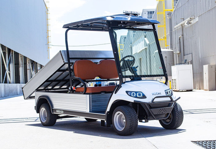ocean cartz three-quarter front view of a white utility electric vehicle with a fully electric dump bed raised for unloading, parked in an industrial area. This specialized electric work cart features a black roof, a black brush guard, and brown two-seater upholstery. The utility golf cart has custom wheels and a clear windshield, with the large aluminum dump bed prominently displayed.
