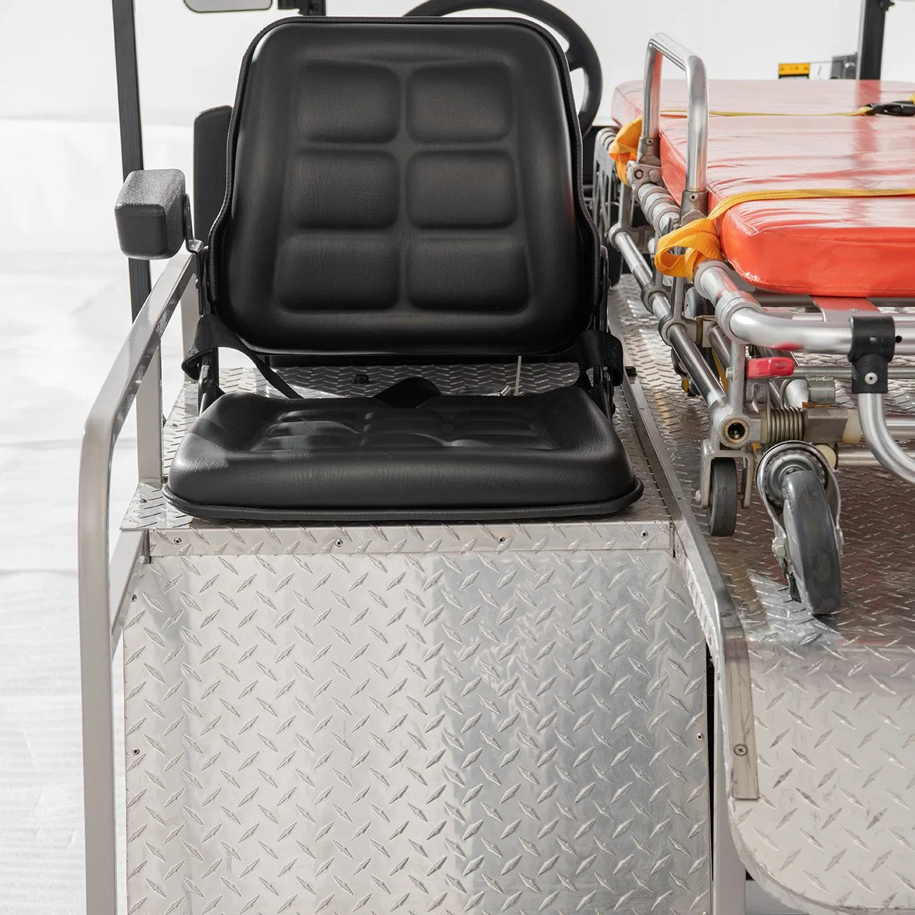ocean cartz close-up detail of the black driver's seat and the diamond-plate metal deck inside a white ambulance golf cart. The utility cart seat is a wide, black, quilted design with armrests. To the right, the rear section of the cart shows the wheels and frame of the orange emergency stretcher, highlighting the specialized, durable construction of the medical utility vehicle.