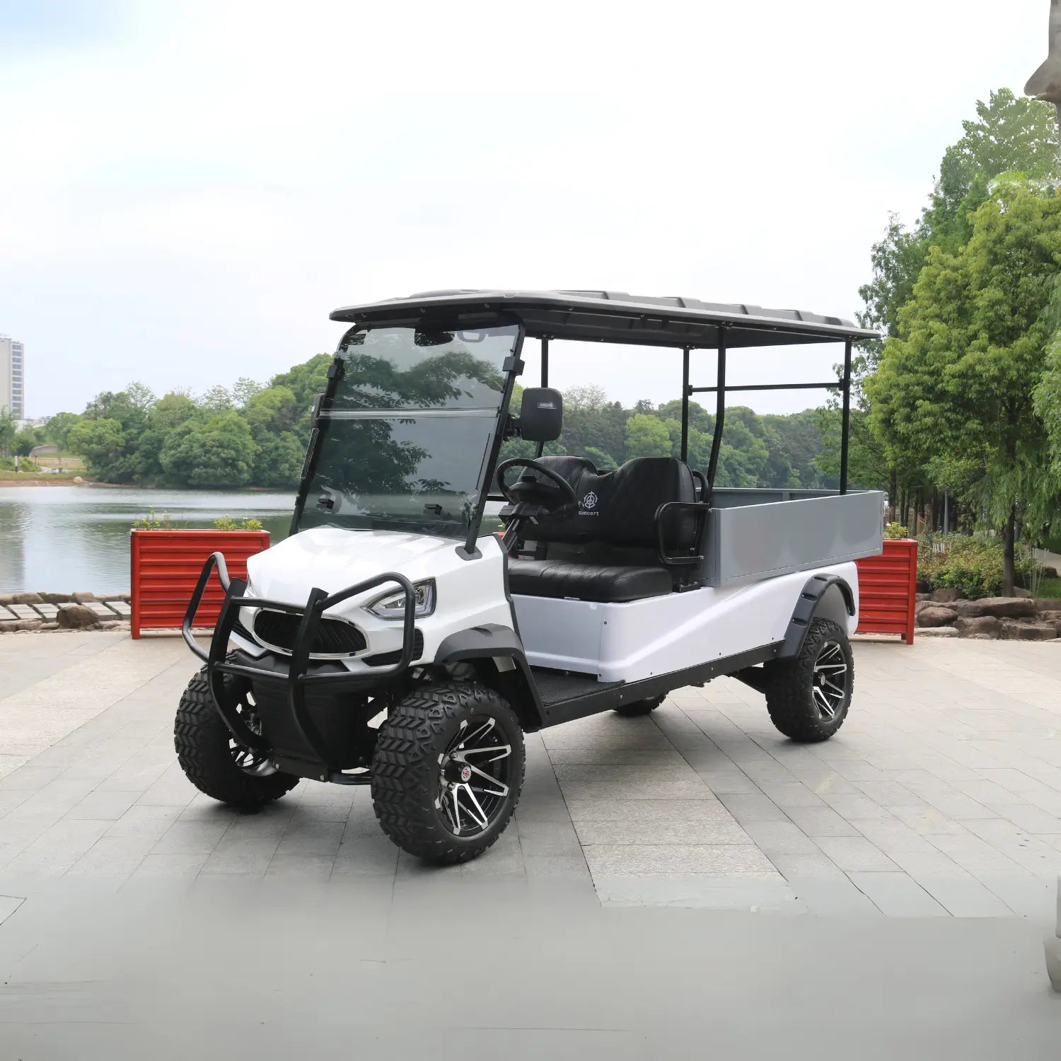 ocean cartz three-quarter front view of a white utility golf cart with a large, grey rear cargo box and a black roof. This off-road electric vehicle is a lifted utility cart featuring black seats, a heavy-duty front brush guard, and aggressive all-terrain tires. It is equipped with custom black and silver wheels and is parked on a paved area next to a body of water with trees and a distant building.
