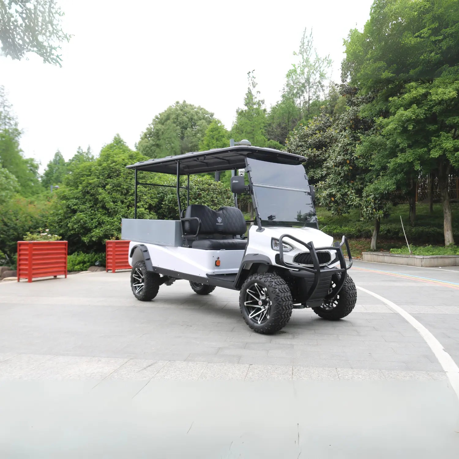ocean cartz side view of a white four-seater utility golf cart with a black roof and a grey rear cargo box. This lifted electric vehicle is an off-road utility cart featuring black seats, a front brush guard, and aggressive all-terrain tires. The cart has custom black and silver wheels and is parked on a paved area surrounded by dense green trees and foliage in a park-like setting.