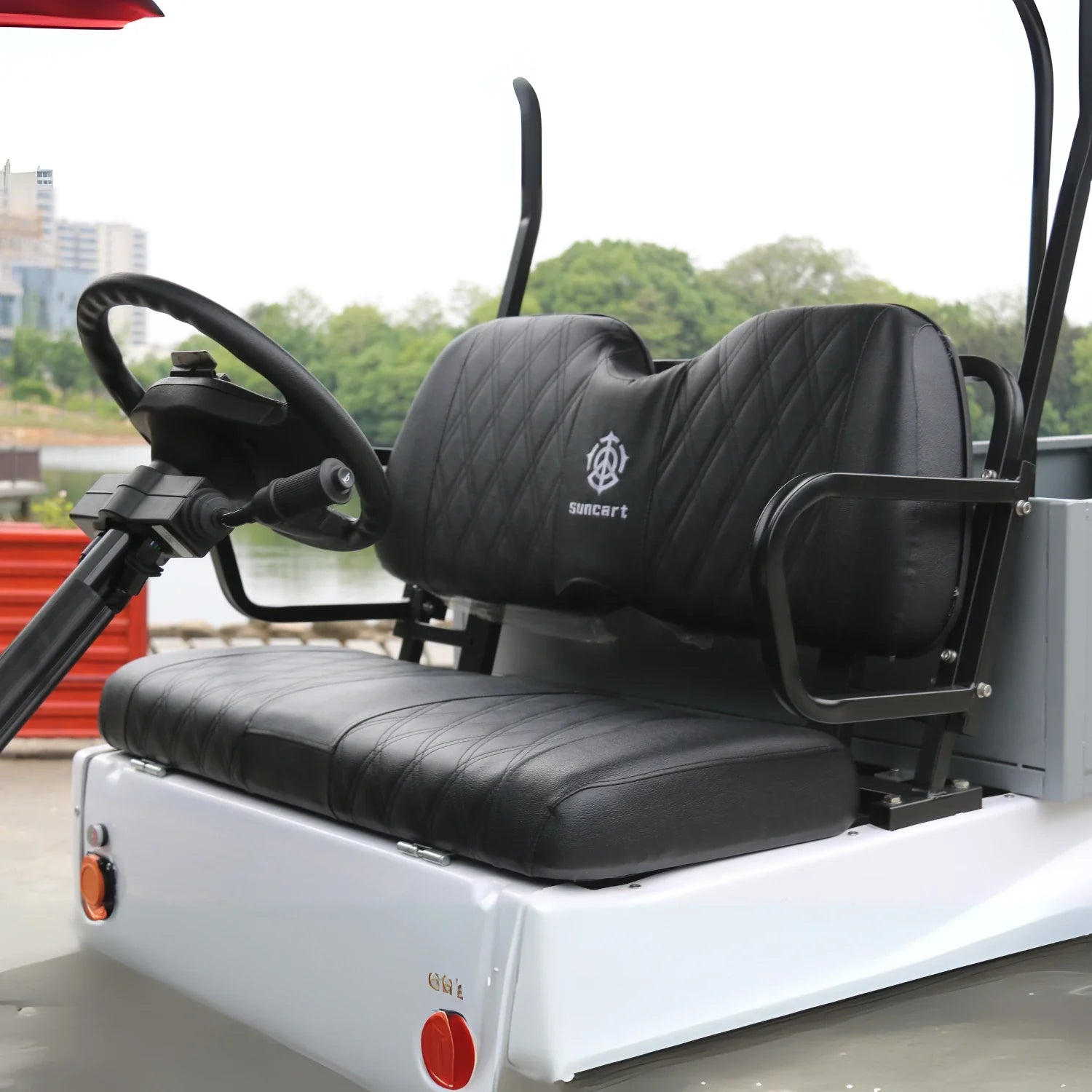 ocean cartz close-up interior view of the black driver and passenger seats in a white utility golf cart. The quilted vinyl seats feature a diamond stitch pattern and a "SUNCART" logo embroidered on the backrest. The image focuses on the luxury seating and the steering column, with the urban outdoor setting, including a body of water and distant buildings, blurred in the background.