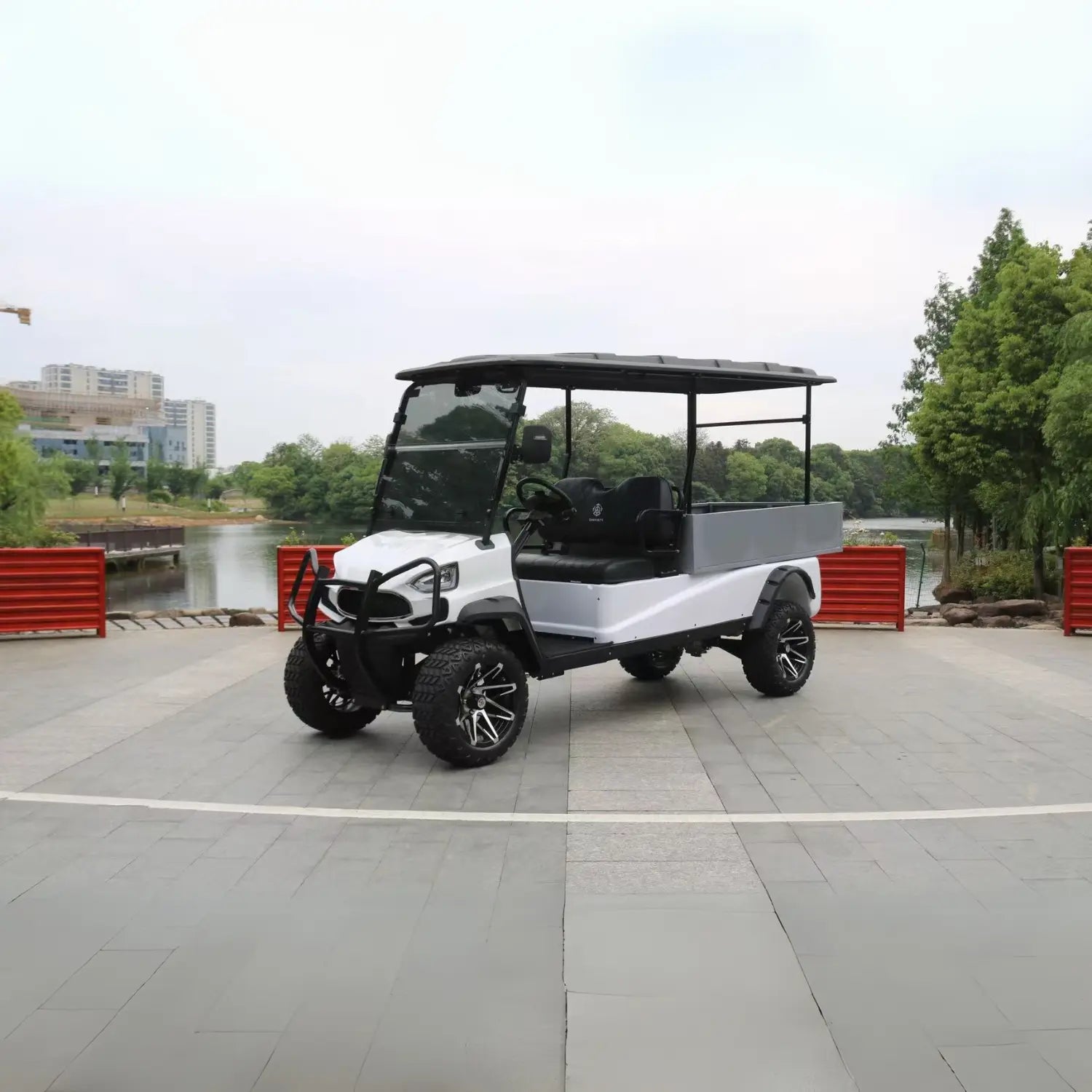 ocean cartz front view of a white utility golf cart with a black roof and a grey rear cargo box, parked on a wide paved plaza. This lifted electric vehicle is an off-road work cart featuring a front brush guard, black seats, and aggressive all-terrain tires. The cart has custom black and silver wheels and is shown near a body of water with trees and city buildings in the background.