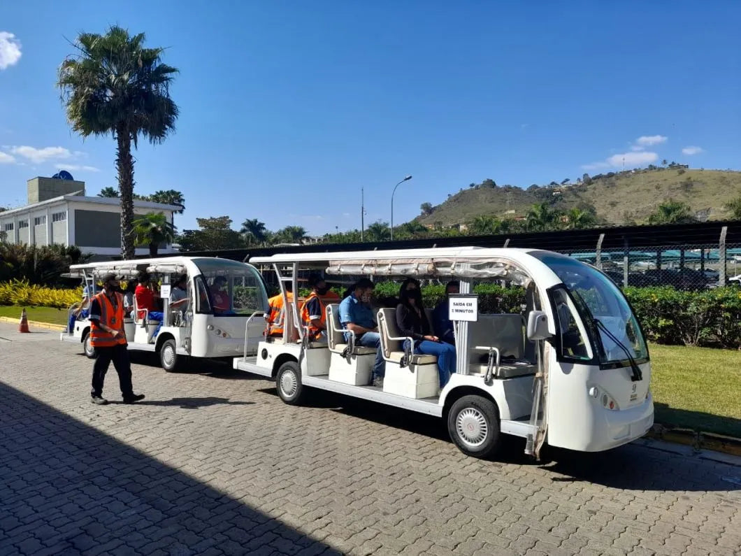 Two white electric tour buses on a paved road with palm trees and buildings in the background.