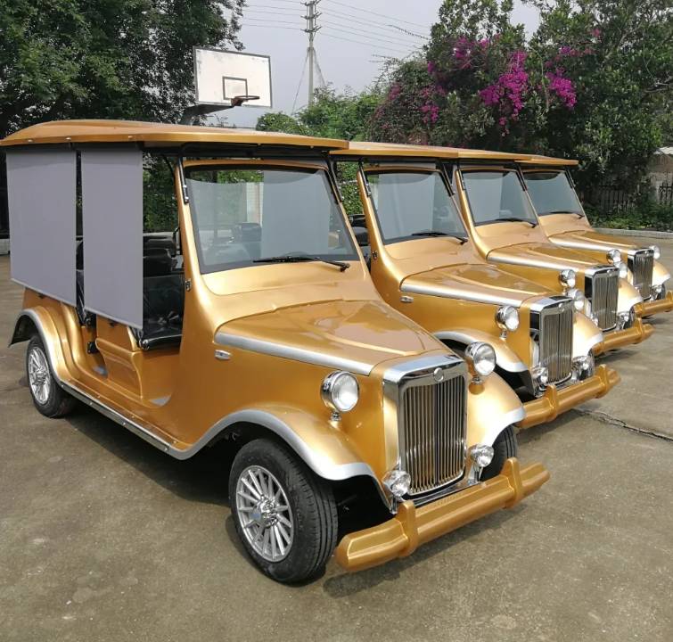ocean cartz three-quarter front view of a line of three gold classic antique-style golf carts with black roofs, parked on a paved court. The front vintage electric vehicle features a gold grille and bumper, silver wheels, and a pulled-down grey side panel for shade. These multi-passenger classic carts are shown in an outdoor setting with a basketball hoop and trees in the background, highlighting their use as resort transport vehicles.