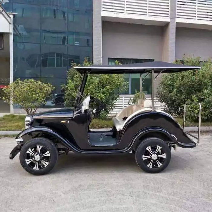 ocean cartz side profile view of a black classic antique-style golf cart with a black roof and large, sweeping fenders. This two-seater vintage electric vehicle features a windshield, light beige or tan upholstery, and black custom wheels with silver accents. The classic golf cart is parked on asphalt outside a modern commercial building with trees and bushes in the foreground.