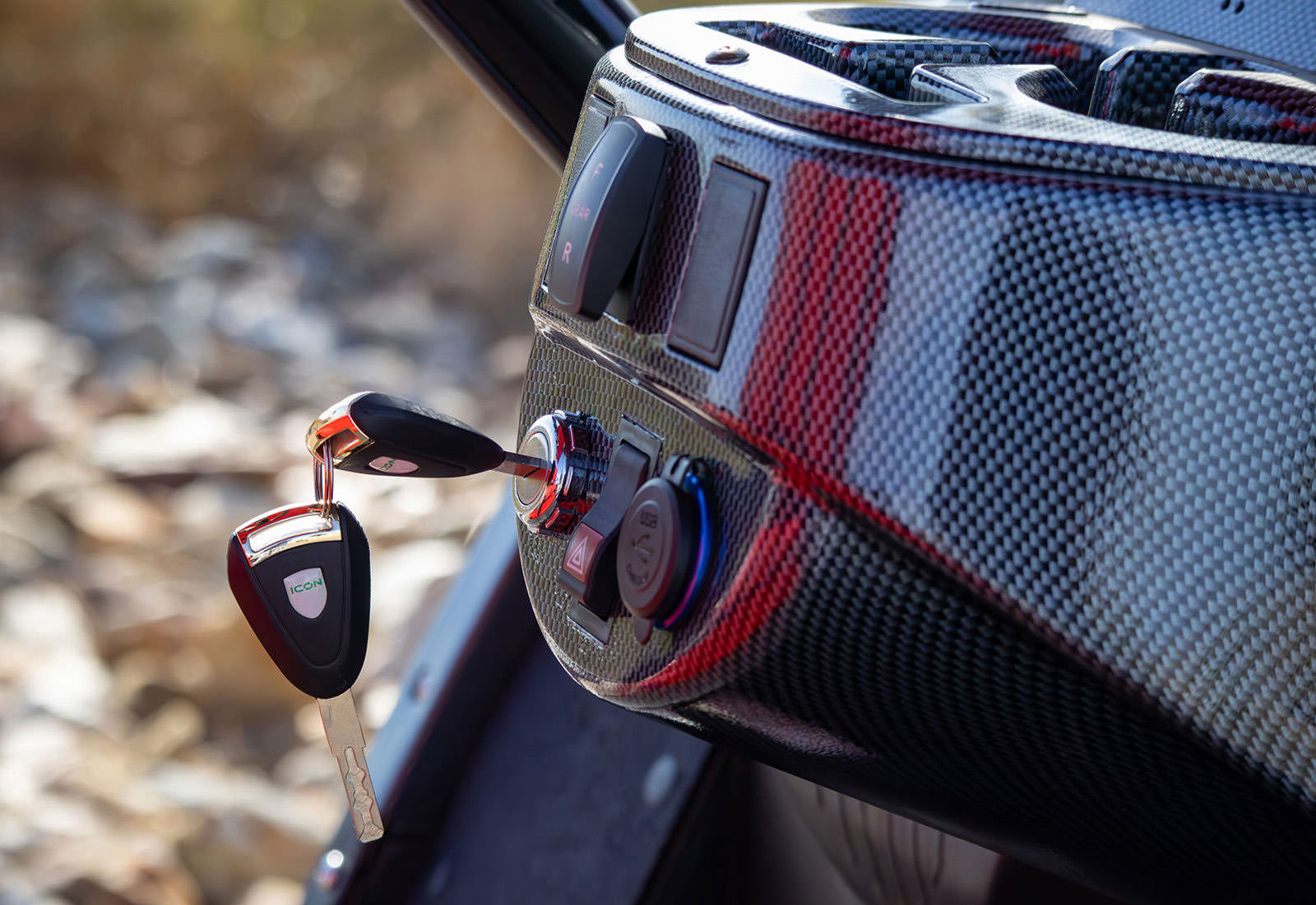 Car key fob attached to a carbon fiber-textured car mirror with a blurred outdoor background.