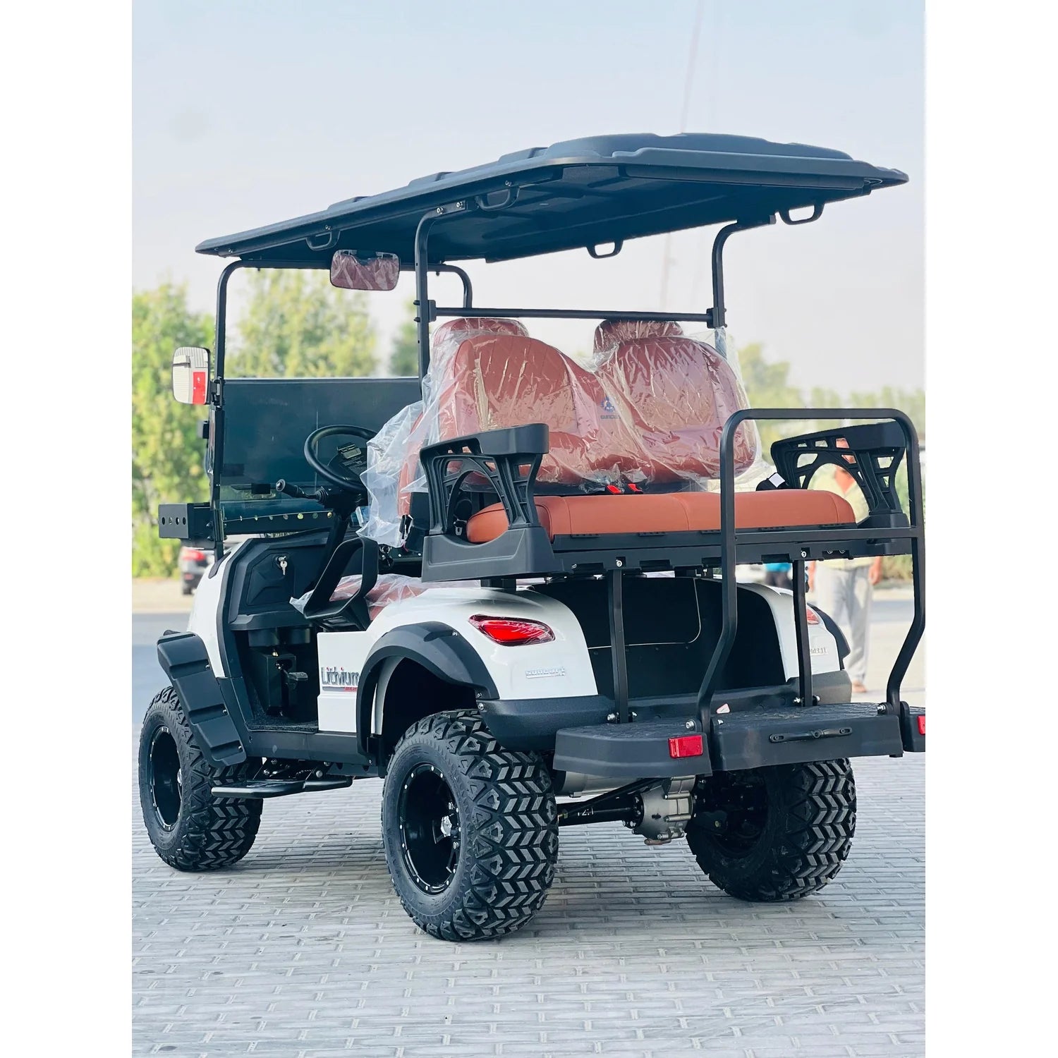 ocean cartz rear three-quarter view of a white lifted golf cart with a black roof and black trim, showing its rear-facing seat. This four-seater utility cart is an off-road electric vehicle with large, knobby all-terrain tires on black custom wheels. It features brown upholstered seats with plastic covers and is parked on a paver walkway next to a street with trees in the distance.