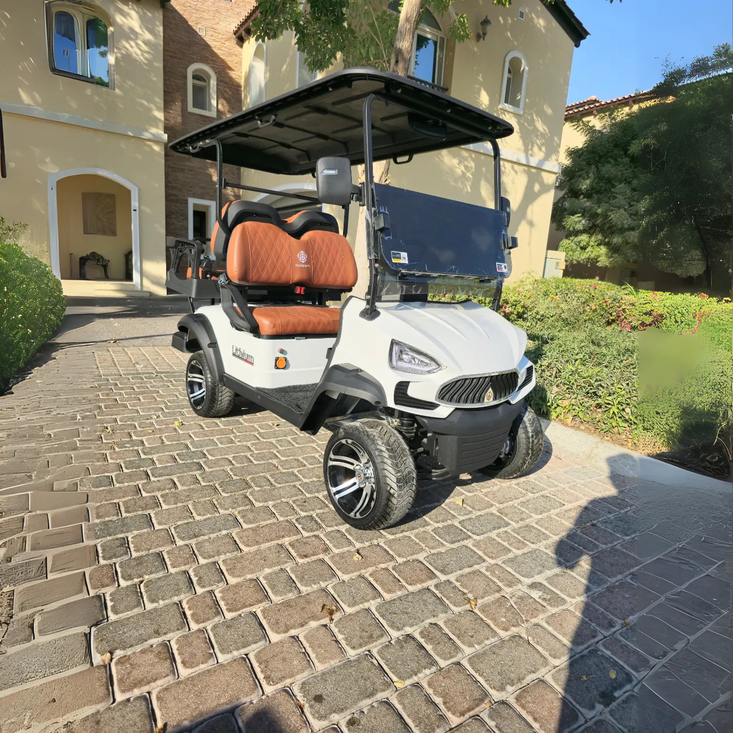 The cart has a modern, aggressive front design, a black roof, and brown, diamond-stitched leather seats with black accents. It is equipped with custom black and chrome wheels and all-terrain tires. The background features a large, Mediterranean-style villa with light-colored stucco and arched windows, surrounded by well-maintained green landscaping. A faint shadow of a person taking the photo is cast on the lower right of the cobblestone surface.