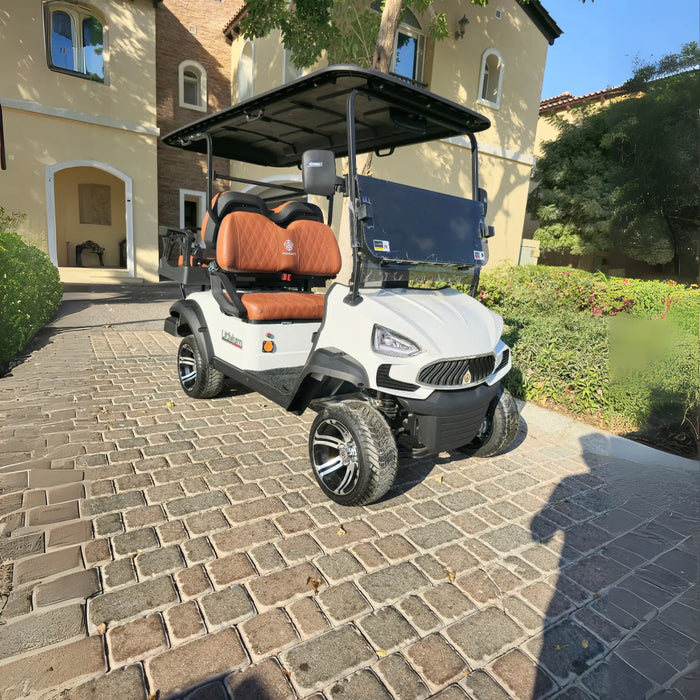 The cart has a modern, aggressive front design, a black roof, and brown, diamond-stitched leather seats with black accents. It is equipped with custom black and chrome wheels and all-terrain tires. The background features a large, Mediterranean-style villa with light-colored stucco and arched windows, surrounded by well-maintained green landscaping. A faint shadow of a person taking the photo is cast on the lower right of the cobblestone surface.