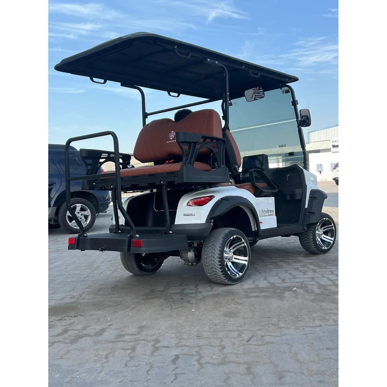 ocean cartz white four-seater luxury golf cart with brown diamond-stitched leather seats and a black roof. This rear three-quarter view highlights the custom chrome wheels and off-road tires, a fold-down rear seat or cargo bed, and a full windshield. The electric vehicle is parked on concrete in an open area with a vehicle and building visible in the background under a light blue sky.