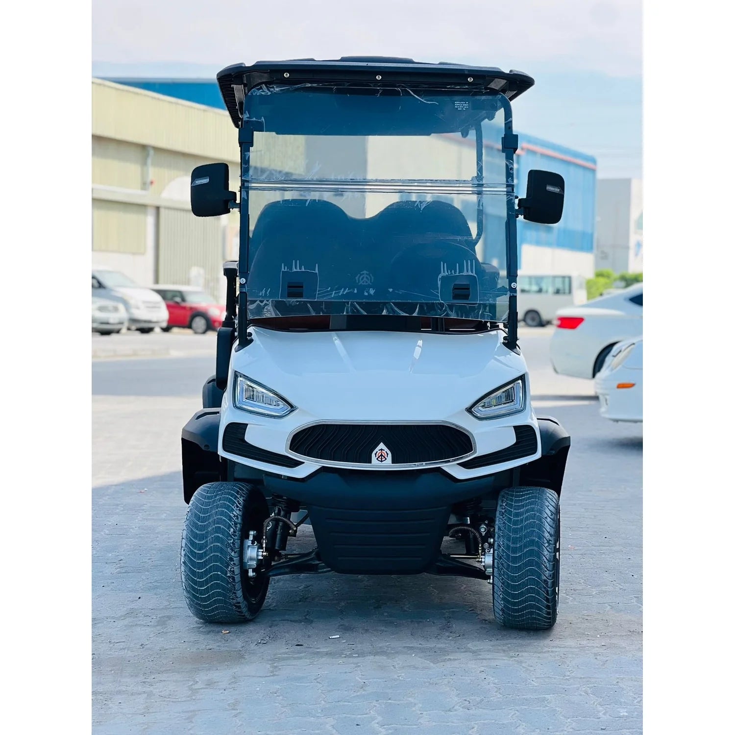 ocean cartz head-on view of a white electric golf cart with a sleek, modern front design, aggressive headlights, and a black grille. The utility vehicle features a clear, tall windshield, side mirrors, and wide, all-terrain tires mounted on black wheels. The off-road golf car is parked in an industrial lot with other vehicles and warehouse-style buildings visible in the background.