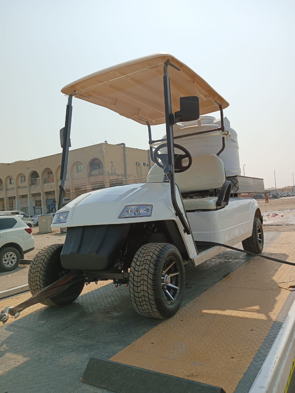 ocean cartz three-quarter front view of a white utility electric vehicle designed as a sprayer or water transport golf cart, secured on the flatbed of a tow truck or trailer. This specialized two-seater work cart features a large white spherical water tank mounted in the rear and a beige roof. The lifted cart is equipped with all-terrain tires and custom black wheels, highlighting its function as a maintenance vehicle being transported in an urban or industrial area.