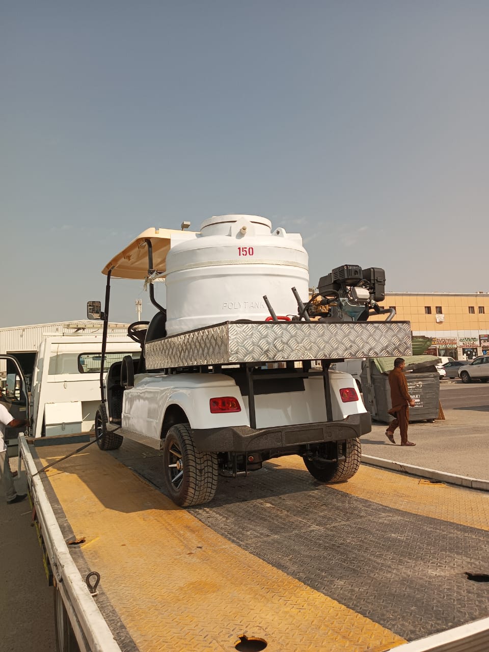 ocean cartz rear three-quarter view of a white utility electric vehicle designed as a sprayer or water transport golf cart, being loaded onto a flatbed tow truck or trailer. This specialized two-seater work cart features a large white cylindrical water tank labeled "150" and "POLY TANK," mounted on a metal diamond-plate platform with a gas-powered pump or engine. The lifted utility cart has a beige roof, all-terrain tires, and custom black wheels, shown in a sunny industrial or commercial area.