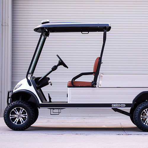 ocean cartz side profile view of a white utility electric vehicle with a black roof and a silver aluminum rear cargo bed, parked in front of a light gray roll-up garage door. This specialized two-seater work cart features brown seats and a lifted suspension. The electric utility golf cart is equipped with all-terrain tires and custom black and silver wheels, highlighting its heavy-duty property maintenance function.