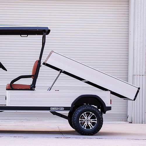 ocean cartz side profile close-up of a white utility electric vehicle demonstrating its fully raised dump bed function, parked in front of an industrial roll-up door. This specialized two-seater electric work cart features brown seats and a silver aluminum rear cargo bed with a hydraulic lifting mechanism clearly visible. The lifted utility golf cart is equipped with all-terrain tires on custom black and silver wheels, highlighting its heavy-duty hauling and dumping capability.