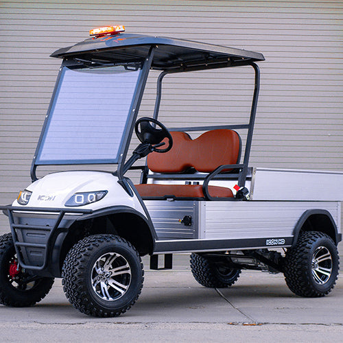 ocean cartz three-quarter front view of a white utility electric vehicle with a black roof and a silver aluminum rear cargo bed, parked in an industrial setting. This specialized two-seater work cart features brown seats, a black brush guard, a large front windshield with a solar panel on it, and an amber emergency light bar on the roof. The lifted utility golf cart is equipped with knobby all-terrain tires on custom black and silver wheels, highlighting its heavy-duty property maintenance function.