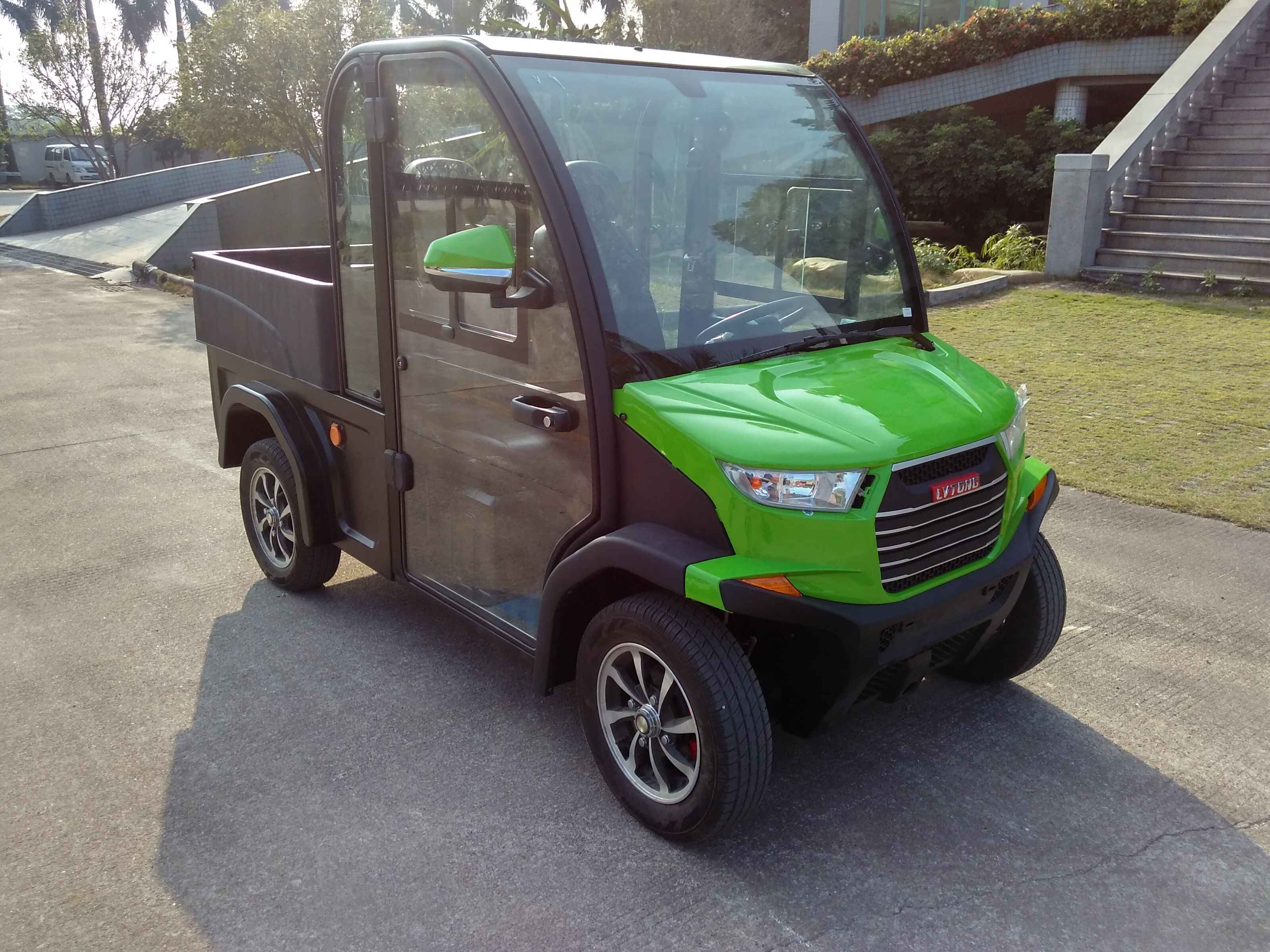 ocean cartz three-quarter front view of a lime green and black utility electric vehicle with a fully enclosed cab and a black rear cargo bed. This two-seater commercial NEV or electric work cart features a bright green hood, black enclosed doors with glass, and a large front windshield. It is equipped with custom black and silver wheels and is parked on an asphalt and concrete area outside a modern building with steps and landscaping, highlighting its use as a compact utility transport vehicle.