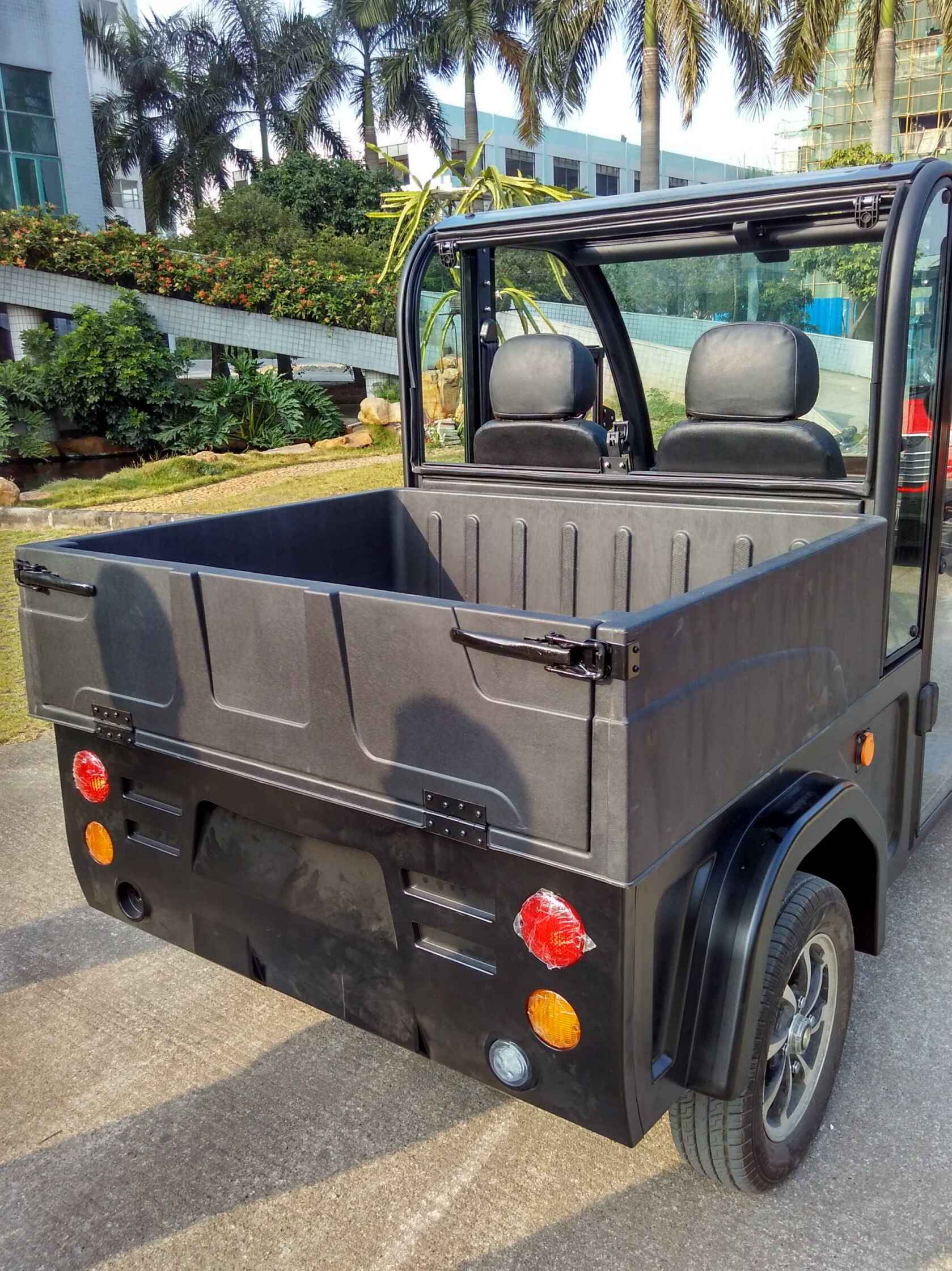ocean cartz rear view of a black electric utility vehicle with a durable, molded plastic cargo box and an open-top cabin, parked outdoors. This two-seater electric work cart features black seats, a clear rear window, and round red and amber taillights. The image highlights the deep hauling capacity of the utility transport vehicle, set in a sunny area with palm trees and a modern building in the background.