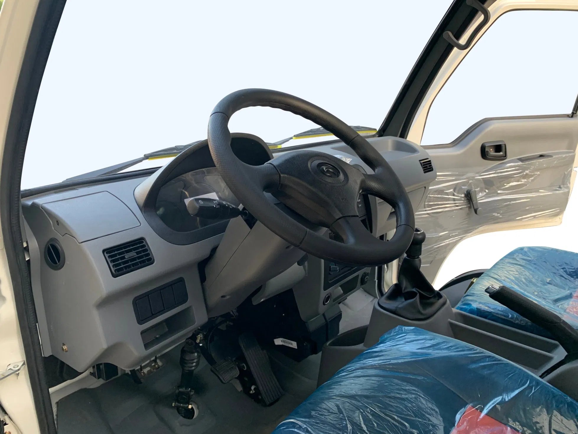 ocean cartz close-up interior view of the driver's cabin in a white electric box truck. The image highlights the grey dashboard, black four-spoke steering wheel, instrument panel, air vents, and manual gear shift lever. The seats are covered in protective plastic, showcasing the simple and functional design of the electric commercial utility vehicle or delivery van.