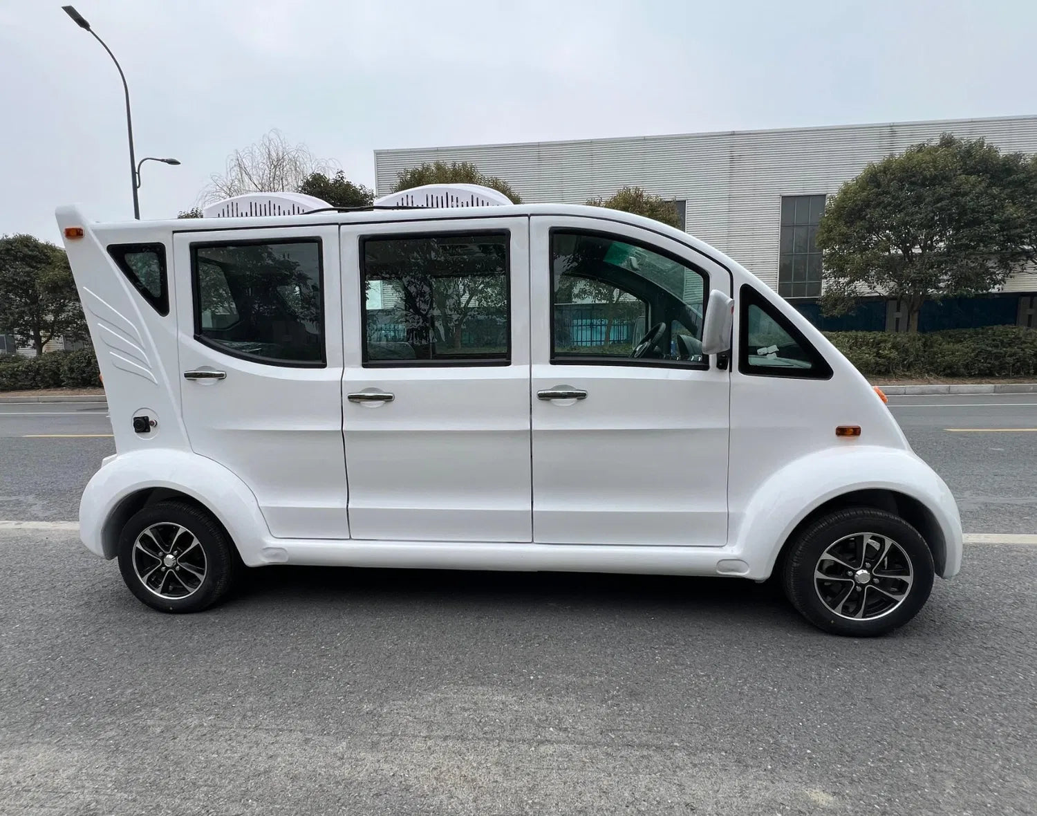 Small white van on a road with a building and trees in the background