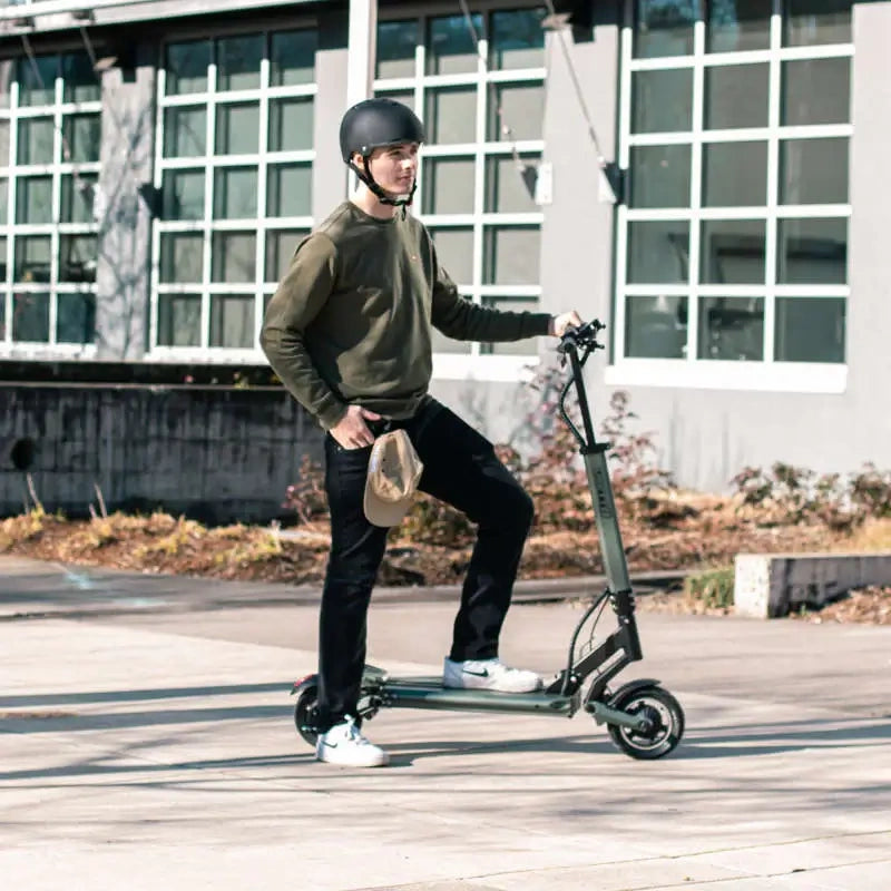 Person riding an electric scooter outdoors on a wooden surface with a building in the background.