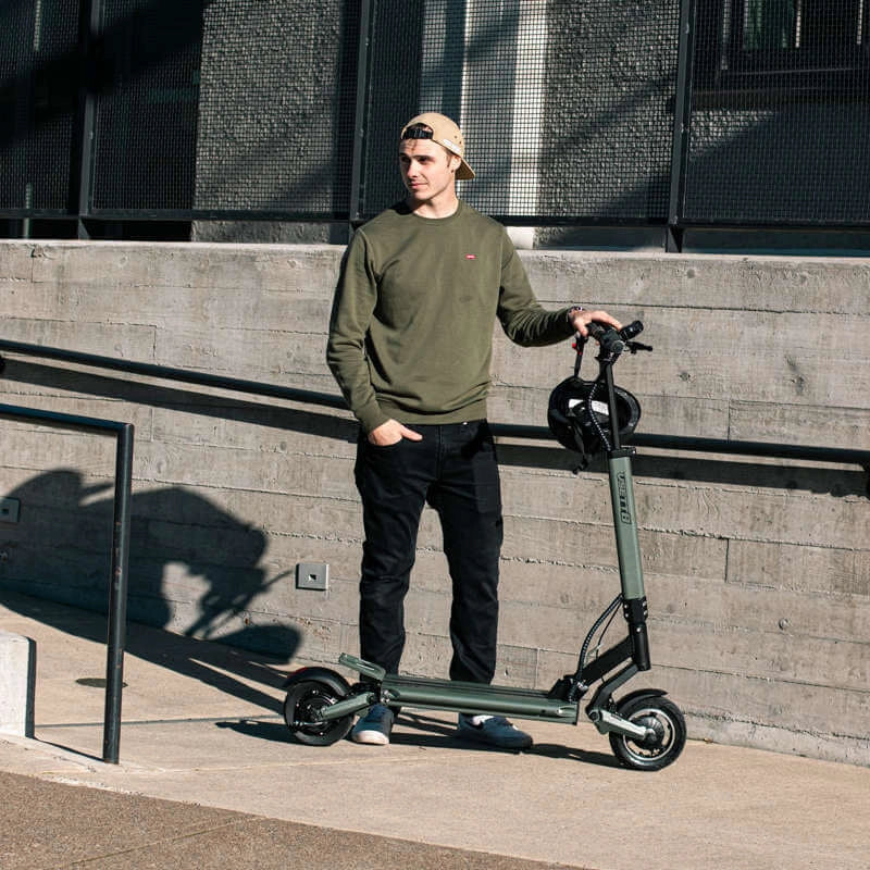 Man standing next to a green electric scooter against a concrete wall.