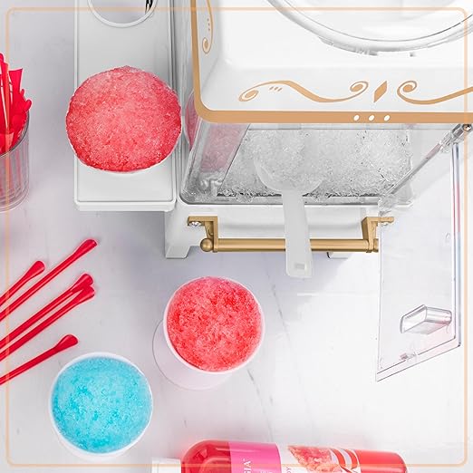 Shaved ice machine with pink and blue shaved ice, red straws, and a bottle of syrup on a white surface.