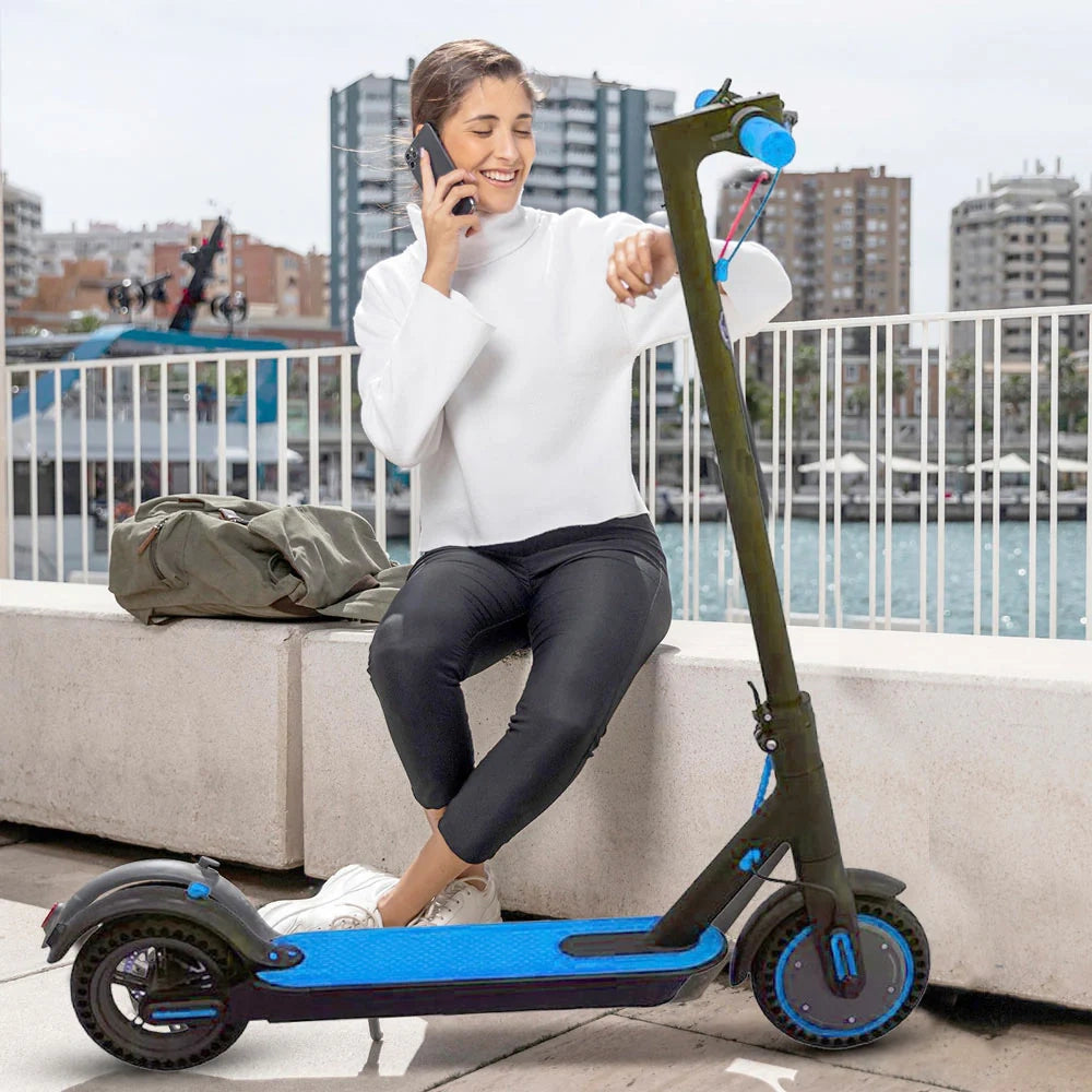 Woman sitting on a blue electric scooter by a waterfront with buildings in the background