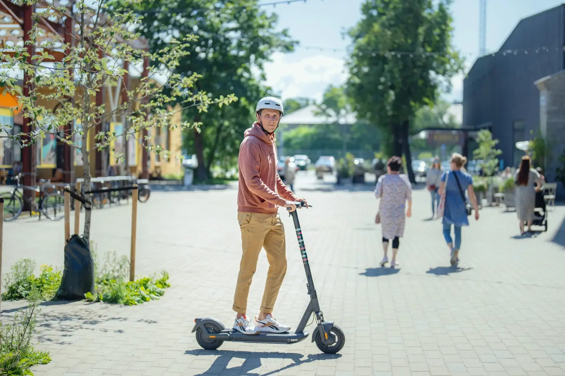 Man riding an electric scooter on a sidewalk with people and vehicles in the background