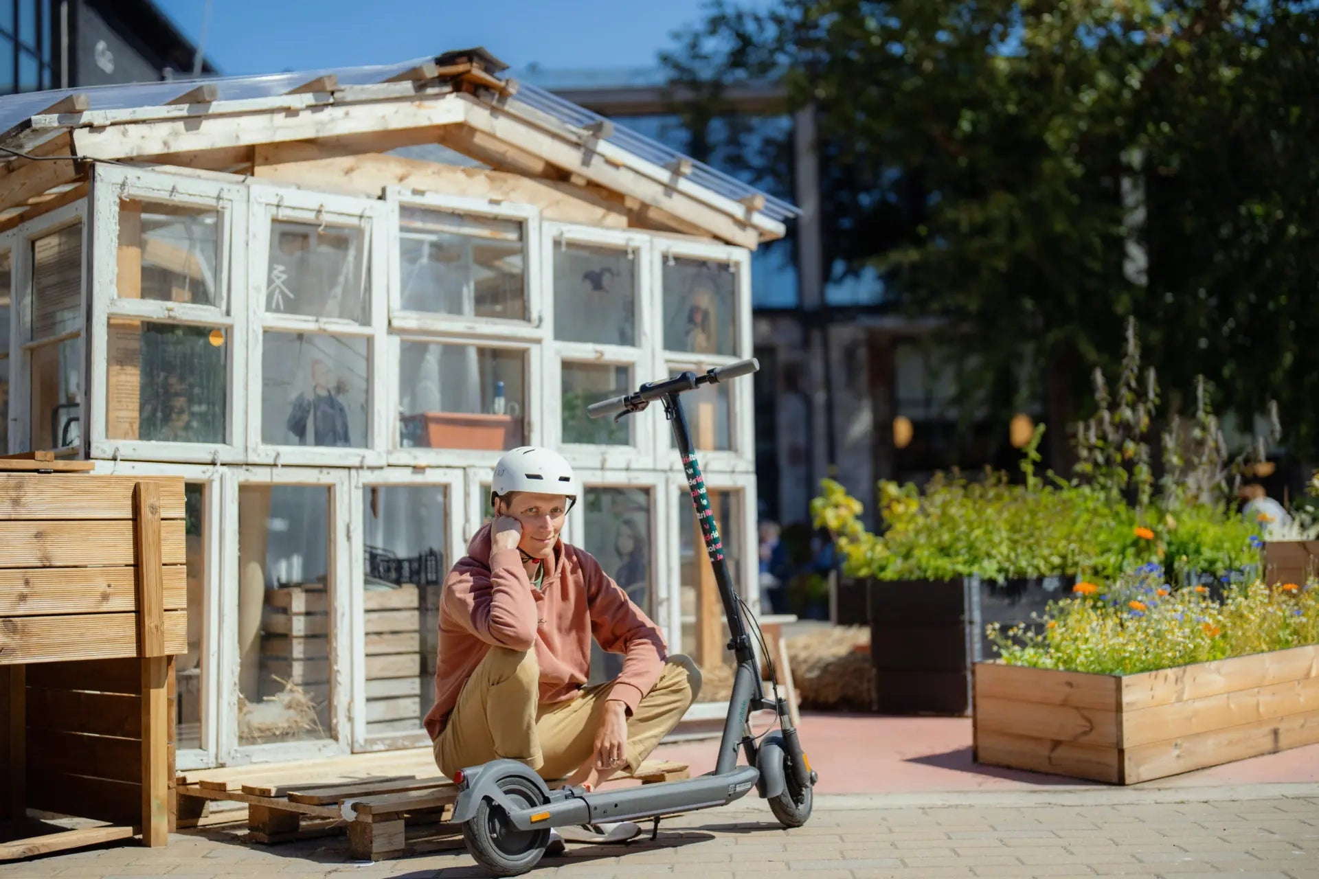 Person sitting on an electric scooter in front of a small wooden greenhouse.