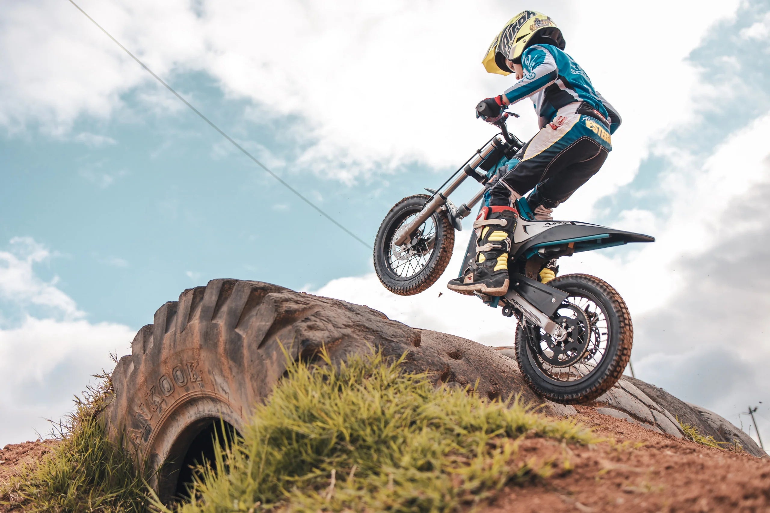 Person riding a dirt bike over a large tire on a track with a cloudy sky.