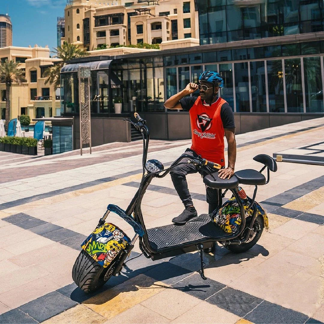 Person sitting on a large black electric scooter with colorful designs in an urban setting.
