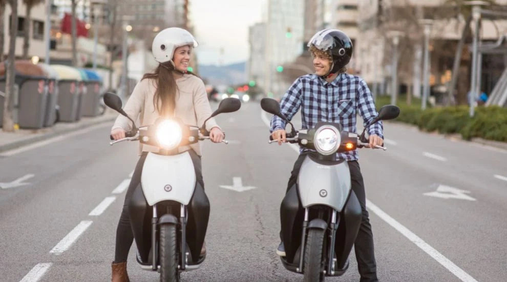 Two people riding scooters on a city street with buildings and traffic lights in the background.