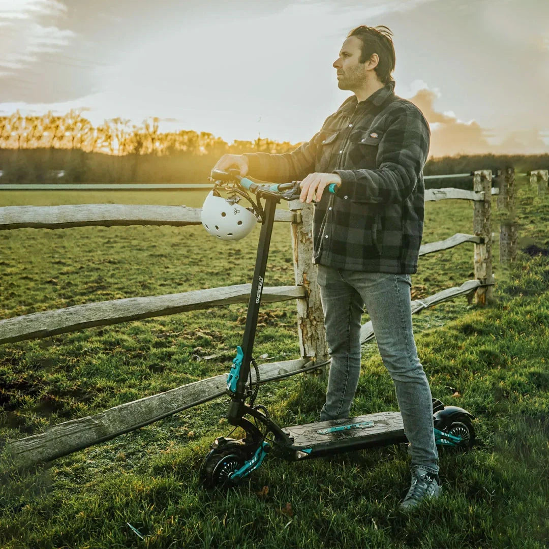 Person standing on a scooter in a field with a wooden fence and sunset in the background