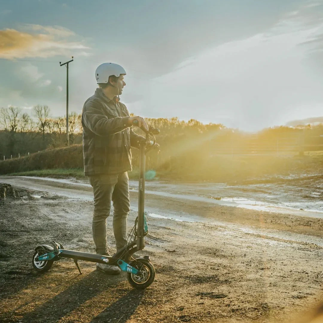 Person with a helmet standing next to an electric scooter on a rural road with a scenic background.