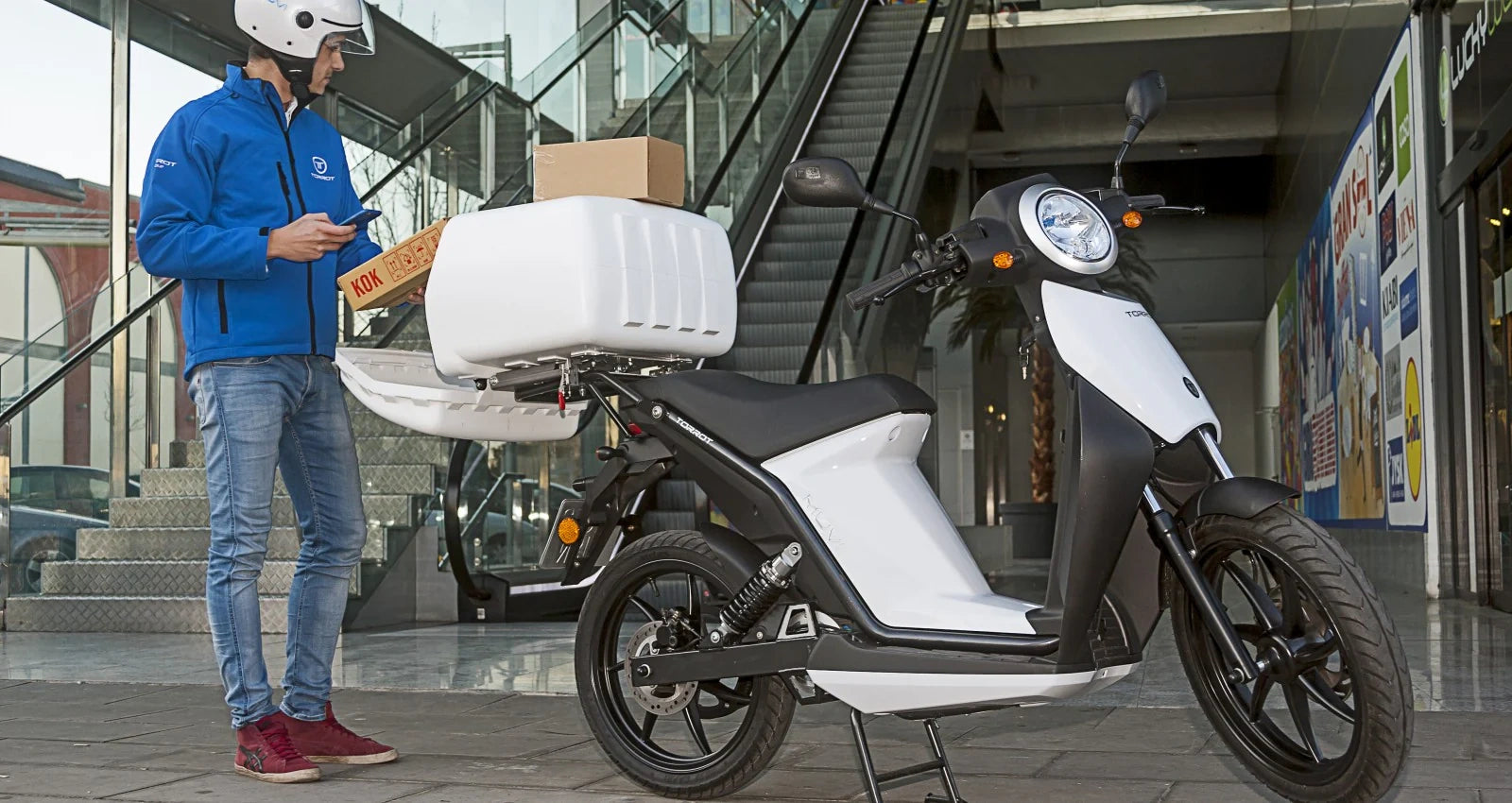 Person with a blue jacket standing next to a white scooter with a cargo box in an urban setting.