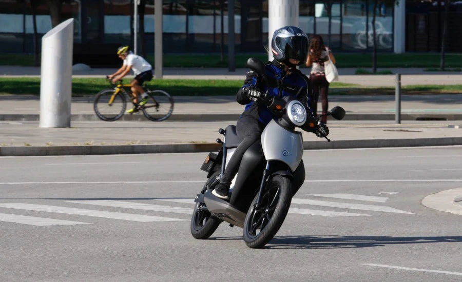 Person riding a scooter on a city street with another person on a bicycle in the background.