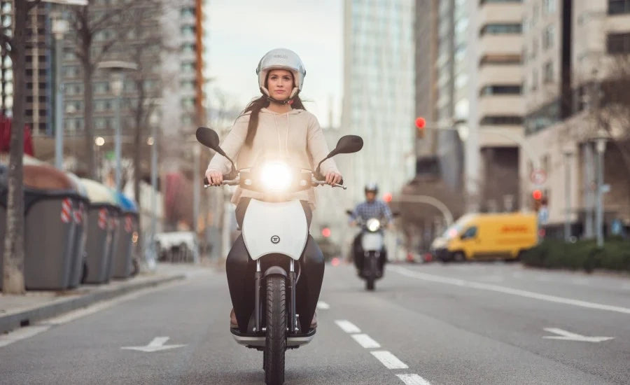 Woman riding a scooter on a city street with buildings and traffic lights in the background