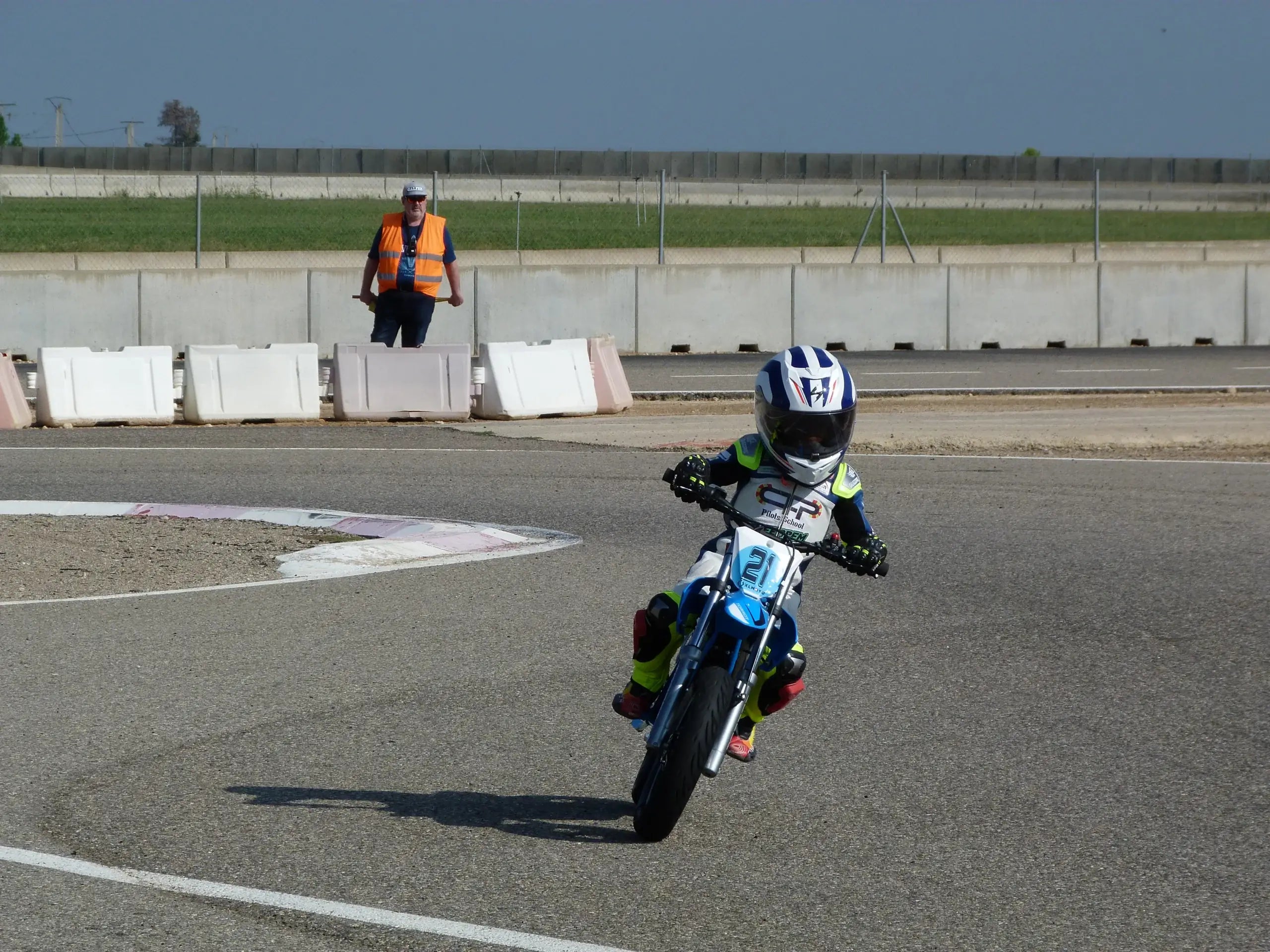 Child on a small motorcycle with a person in an orange vest in the background on a track.